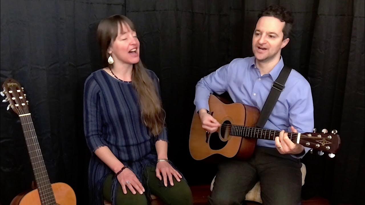 A man and woman are seated, playing acoustic guitars in front of a black curtain backdrop. Aliped Productions Footage rich...